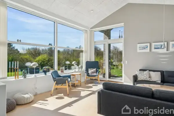 En lys stue med store vinduer, der giver udsigt til en grøn have. To stole og et bord står ved vinduet. På væggen hænger kunstværker over en sofa.