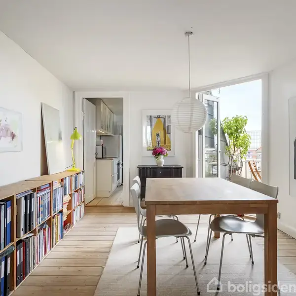 Spisebord omgivet af stole i en lys spisestue. Til højre ses en åben altandør med en plante udenfor. Væggene har kunst og reoler fyldt med bøger.