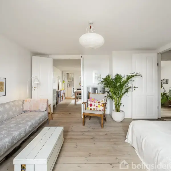 En lys stue indeholder en grå sofa med mønster, en hvid sengebord, og en hvid loftslampe. En stor potteplante står ved siden af en farverig stol, omgivet af hvide vægge med kunst.