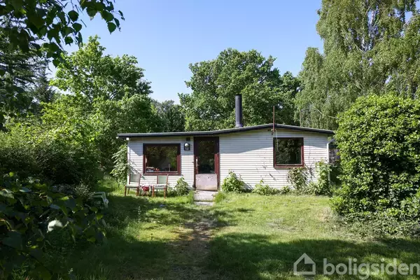 Et lille hus står i en frodig have, omgivet af grønne træer og buske. To vinduer og en dør er synlige på facaden.