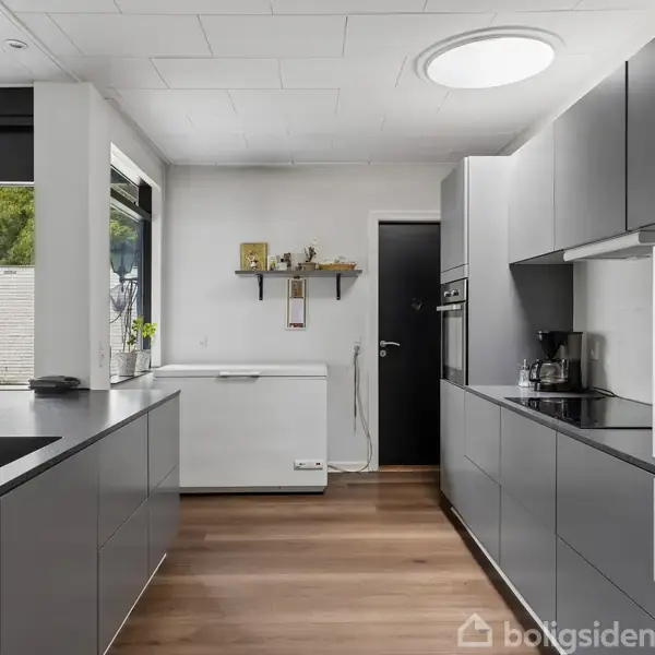 Et moderne køkken med grå skabsfronter, sort bordplade og indbyggede hvidevarer. Rummet har et stort vindue, en hylde med dekorationer, og en trægulvbelægning, hvilket giver en lys atmosfære.