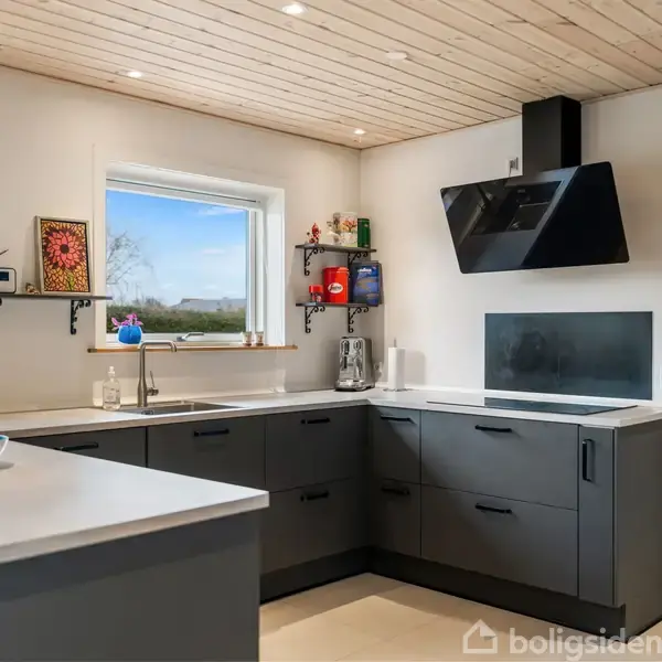 Køkken med mørkegrå skabe og sorte emhætte; står i hjørnet med to vinduer. Lyse trælofter og hvide vægge skaber en lys atmosfære. Bordpladen er lys, og der er en frugtskål.