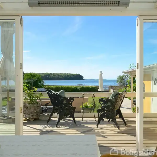 To sorte stole står på en terrasse med udsigt til havet og en grøn ø i det fjerne. Omgivelserne er rolige, med klart blåt hav og himmel.