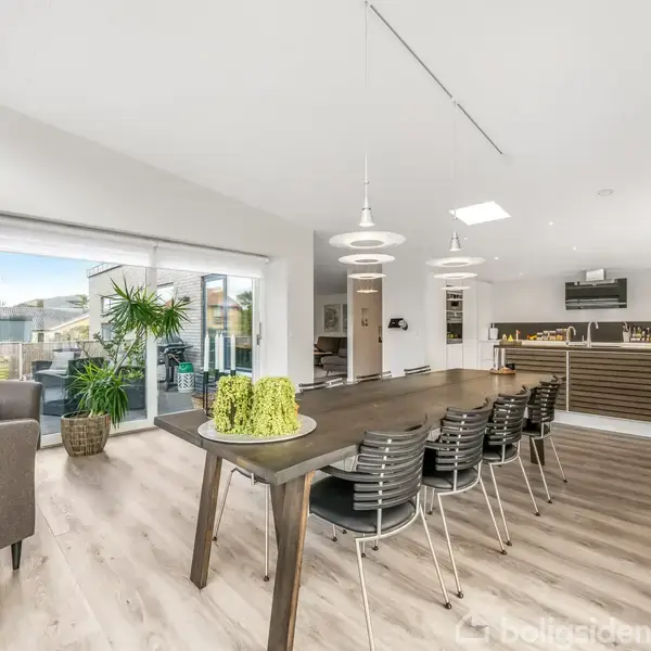 En moderne spisestue med et langt spisebord og stole omkring. Der er grønne planter ved siden af store vinduer, der åbner op til en terrasse. Lyse trægulve, enkelt indretning.