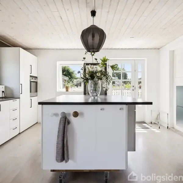 Køkkenø med blomsteret vase står i midten, omgivet af hvidt køkkenskabe og lysindfald fra store vinduer. En gråt håndklæde hænger fra øen, og en moderne lampe hænger over.