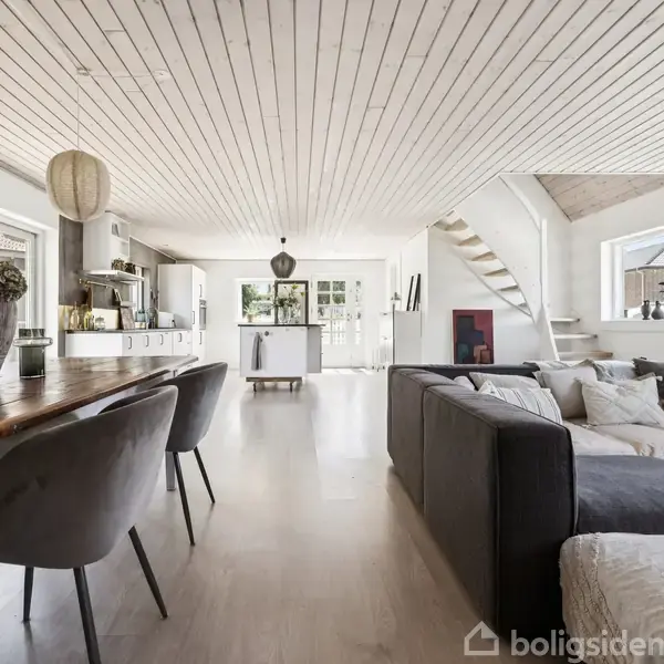 Stue med spisebord og stole i forgrunden, åben til et køkken med moderne apparater. Et sort hjørnesofa står ved vinduer, og en trappe leder til øverste etage. Ikke noget tekst.