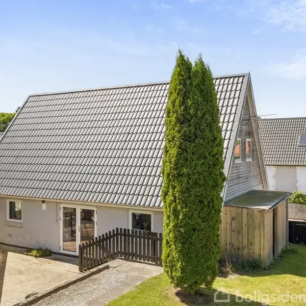 Et hvidt hus med skråt tag står omgivet af en have med græs og få træer. Solen skinner, og huset har en træterrasse med et sort hegn.