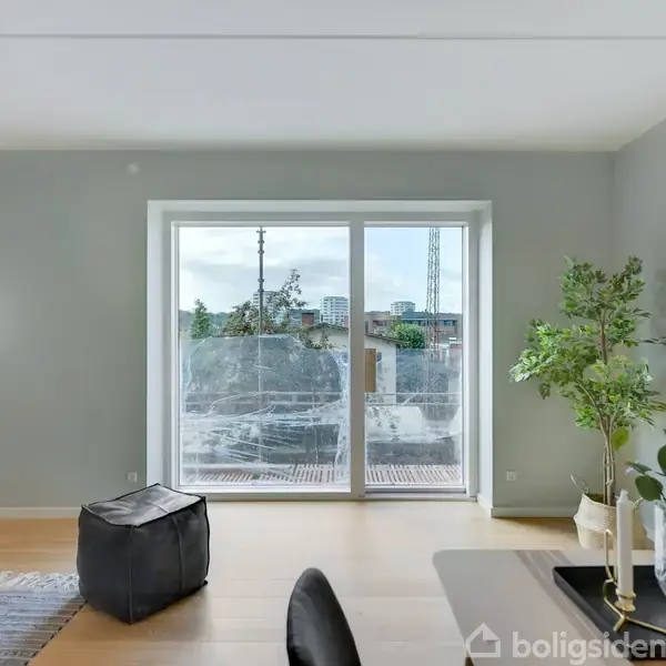 Stue med moderne indretning; lys flyder ind gennem store vinduer, der fører til en balkon. Rummet indeholder en puf, planter, et rundt bord og en hængende lampe.