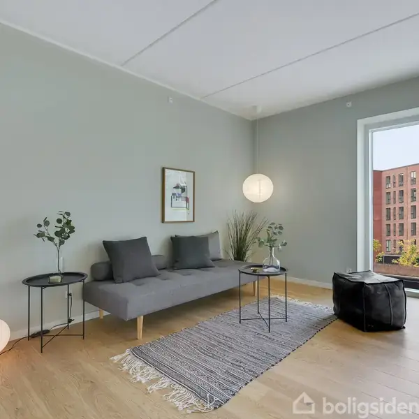 Grå sofa står på tæppe, omgivet af små borde med planter. En stor rude viser en moderne bygning udenfor. Enkelt stuelampe hænger i hjørnet, der skaber en rolig atmosfære.