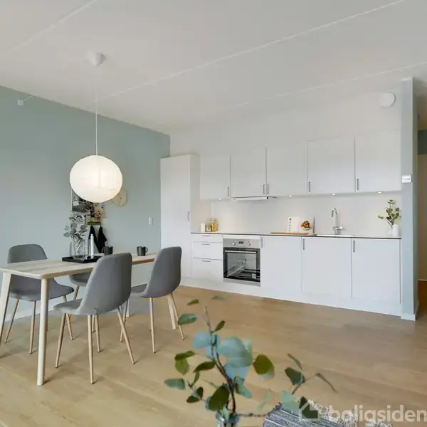 Spisebord med fire stole står i en moderne spisestue. Lys kuglelampes lyser over bordet. Køkken med hvide skabe og integreret ovn i baggrunden. Trægulv og lyseblå vægge.