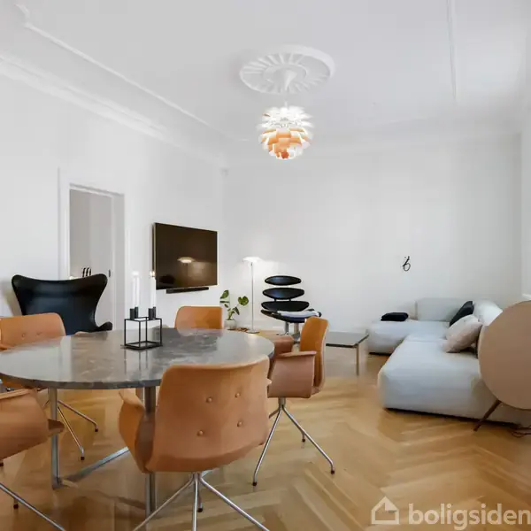 Et rundt marmorbord med brune stole står i en moderne stue. En lys sofa og stilfulde stole står ved tv'et. Rummet har trægulv og hvidmalede vægge.