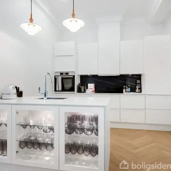 Moderne, hvidt køkken viser en ø med indbygget opvaskemaskine og glashylder fyldt med glas. To hængelamper lyser rummet op. Baggrunden har indbygget ovn og sort backsplash.