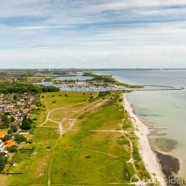 Strand grænser op til en grøn mark, der strækker sig mod et boligområde med huse. Havet er roligt, og himlen er delvist overskyet.