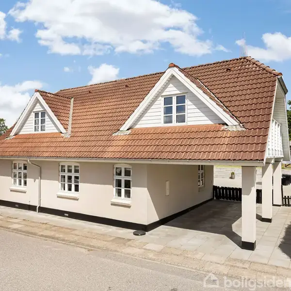 En hvid, étplans bolig med rødt tegltag står i et boligkvarter, omgivet af en lav hæk. Der er en carport til højre, og himlen er klar med nogle skyer.