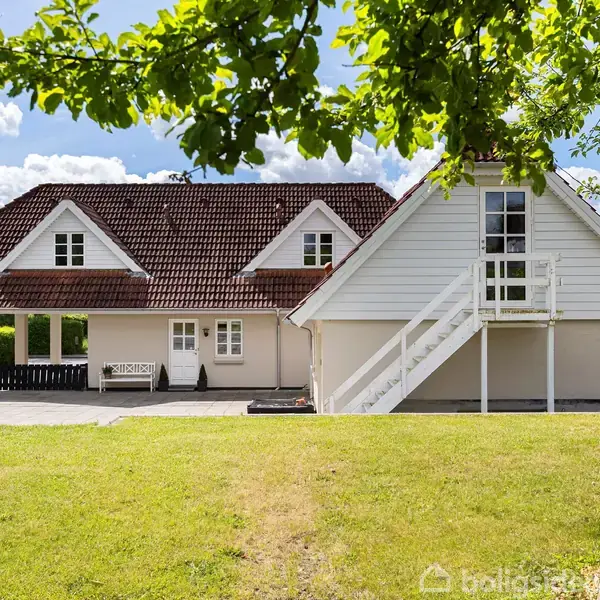 Et hvidt hus med rødt tegltag har en udendørs trappe og altan. Det står i en solrig have med blå himmel og grønne træer omkring.