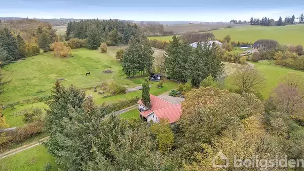 Et rødt taghus omgivet af træer ligger i en frodig, grøn landskab. Bagved er der en mark med en hest, og skoven strækker sig til horisonten under en klar himmel.
