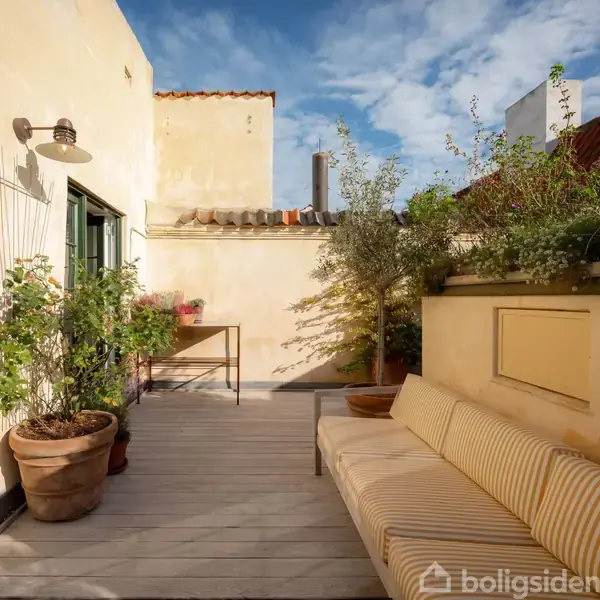 Terrasse med stribet sofa langs en væg, planter i potter omkring, og et bord i baggrunden. Sollys falder på de beige vægge, mens himlen har lette skyer.