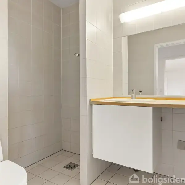 Toilet i et badeværelse, står ved siden af en brusekabine. Hvide fliser dækker væggene, mens en spejlvæg med lys hænger over en vask. Ingen tekst udover "boligsiden".