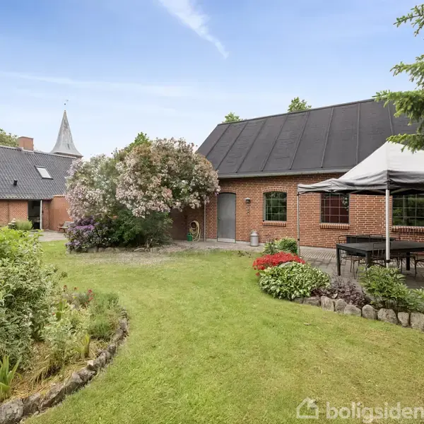 Rødstensbygning med mørkt tag står stille i en frodig have. En pavillon med bord og stole står til højre. Flere buske og blomster omgiver bygningen.