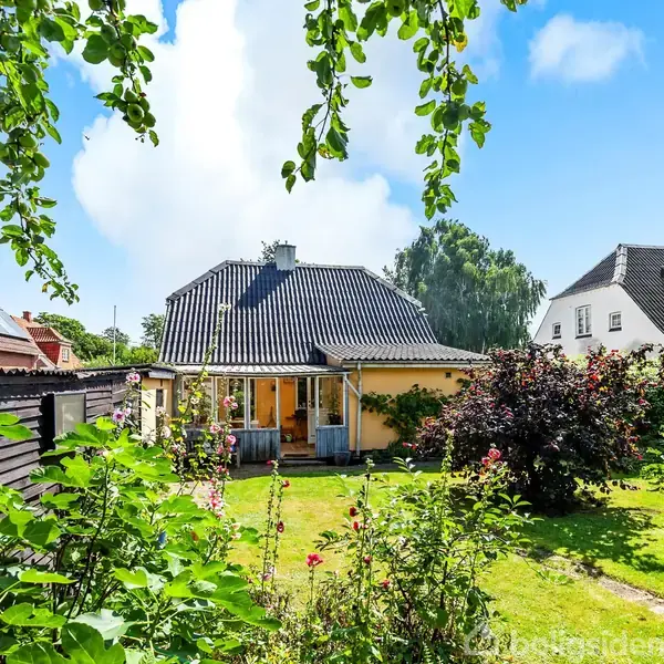 Et gult hus med mørkt tag står omgivet af en frodig have med buske og træer i et boligområde. Solen skinner på den grønne græsplæne.