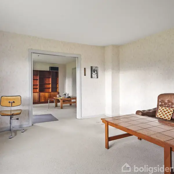 Brunt sofabord står i stue med hvidt tæppe. Ved siden af er en brun lædersofa og en kontorstol. Bagved ses et værelse med bogreoler og træmøbler.