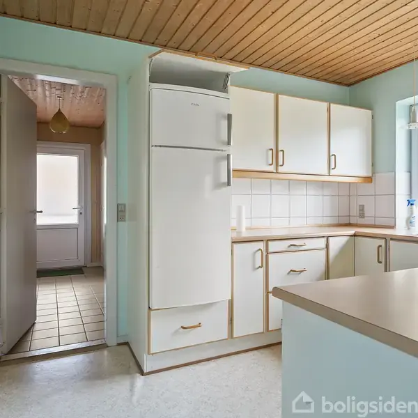 Køkken med hvide skabe og køleskab, hvor lys strømmer ind gennem et stort vindue. En åben dør fører til en lysfyldt gang med træloft.