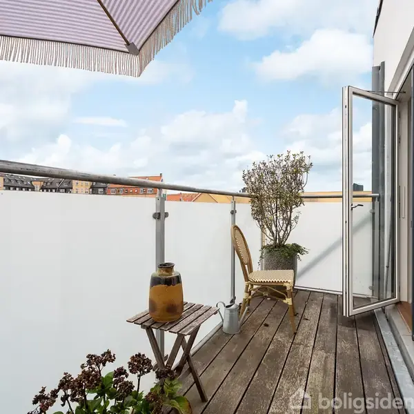 Parasol står over en trædækket altan med en stol, bord og planter. Altandøren står åben. Udsigten viser en blå himmel med skyer og bytag i baggrunden.