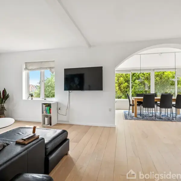 Sort lædersofa i en moderne stue, hvor et vægmonteret TV hænger. Stuen åbner op til en spisestue med et stort vinduesparti, der viser frodig natur udenfor.