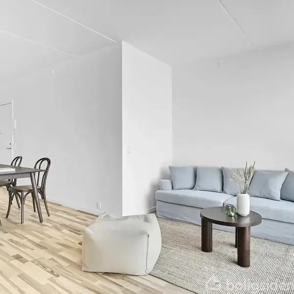 Sofa står i en lys stue ved siden af et lille bord med en vase. I baggrunden ses et spisebord med stole på trægulv.