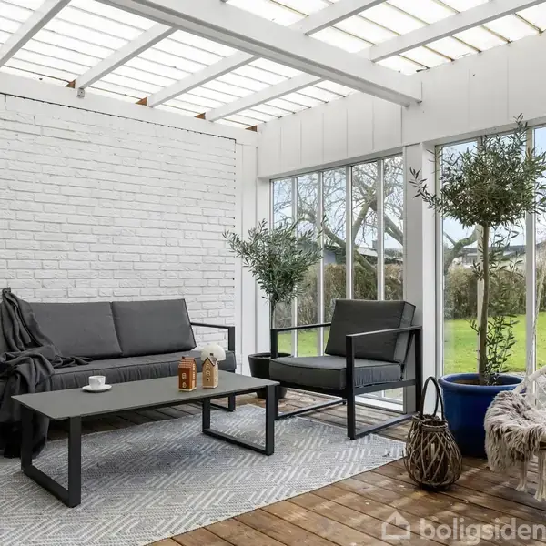 Sofasæt med gråt betræk står på et tæppe i et lyst udestueområde. En hvid hængestol hænger til højre, omgivet af grønne planter og store vinduespartier med udsigt til haven.