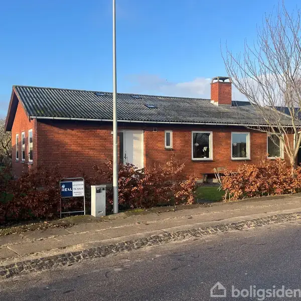 Rødt murstenshus med sort tag står stille i en forstadsgade med en lyseblå himmel. Et til salg-skilt fra "Realmæglerne" er placeret foran huset.