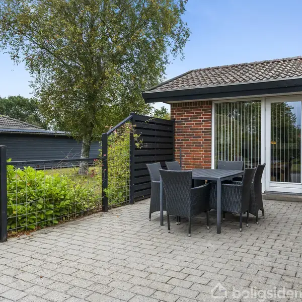 Terrasse med firkantet bord og seks stole på flisebelagt område ved hus med røde mursten og hvide vinduer. Omgivet af hæk og træer, der skaber en hyggelig atmosfære.