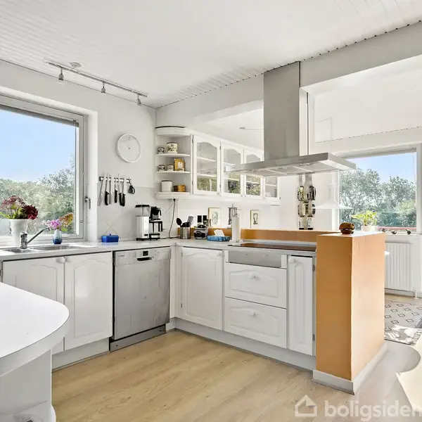 Et hvidt køkken med åbne hylder og en ovnø. Det er veloplyst af store vinduer, der fører ud til en terrasse med udsigt over grønne træer.