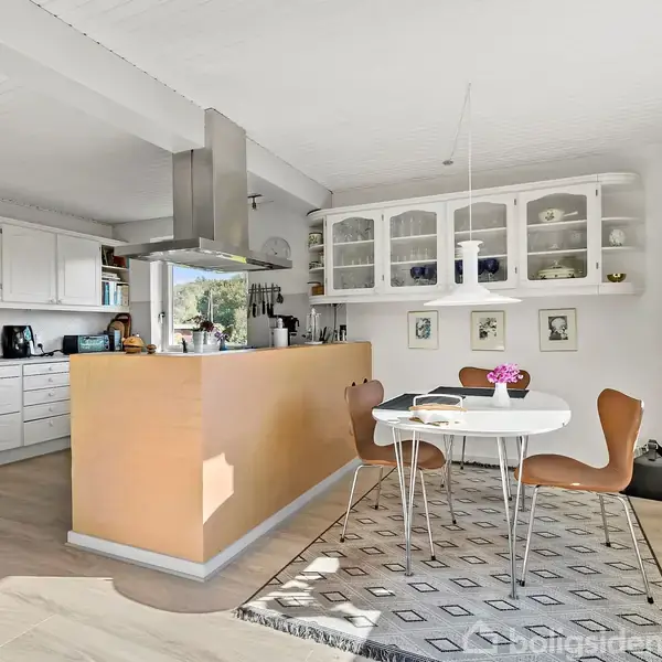 Et spisebord med blomster og tre stole står på et mønstret tæppe ved siden af et lyst køkken. Skabe med glaslåger og en emhætte er i baggrunden.