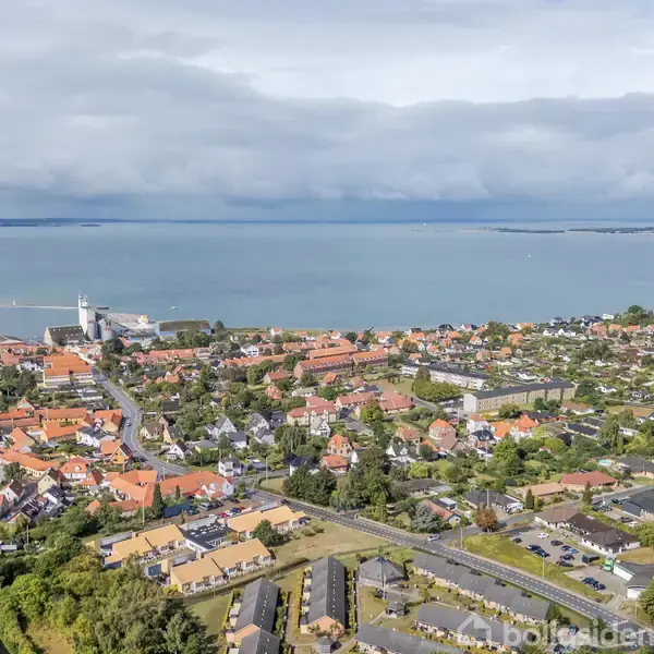 By indeholdende røde taghuse og en stor kirke nær kystlinjen. Huse strækker sig mod havet under en skyet himmel, med grønne områder spredt omkring. Kysten møder vandet i baggrunden.