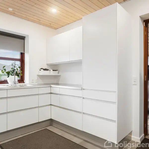 Et køkken med hvide, moderne skabe og skuffer. En plante står i vindueskarmen. Træloft og lys trægulv skaber en lys atmosfære. Døren fører ud til en terrasse.