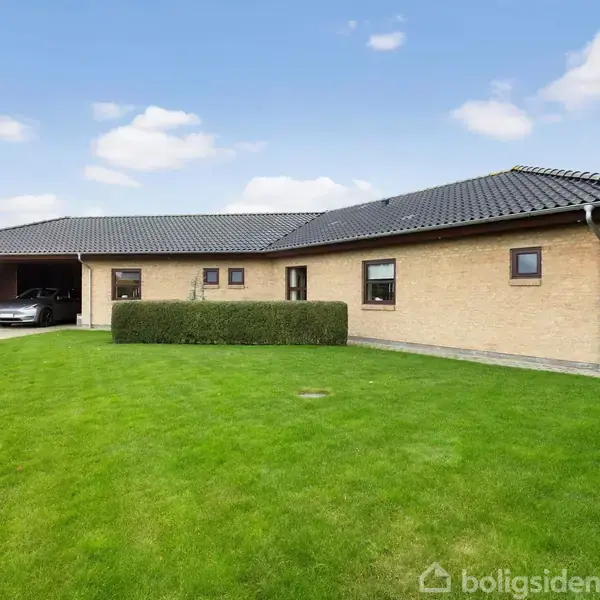 Et moderne, enfamilieshus i gule mursten står stille på en velplejet græsplæne. En bil er parkeret i en overdækket garage til venstre. Himlen er klar med få skyer.