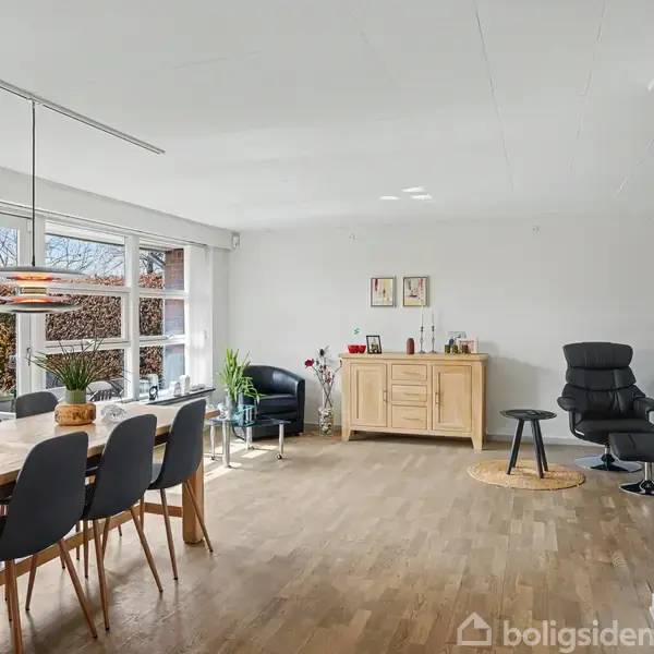 Langbord omgivet af sorte stole står i en lys stue. Et skab med dekorationer står til højre, og en sort lænestol står i hjørnet. Store vinduer giver udsigt til have.
