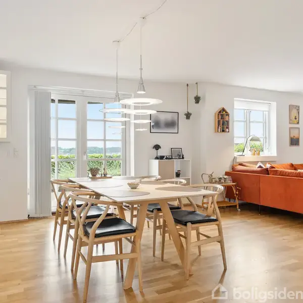 Spisebord med seks stole står i et lyst rum med trægulve. Store vinduer lader naturligt lys ind, og en orange sofa giver kontrast. Planter og kunst pryder rummet.