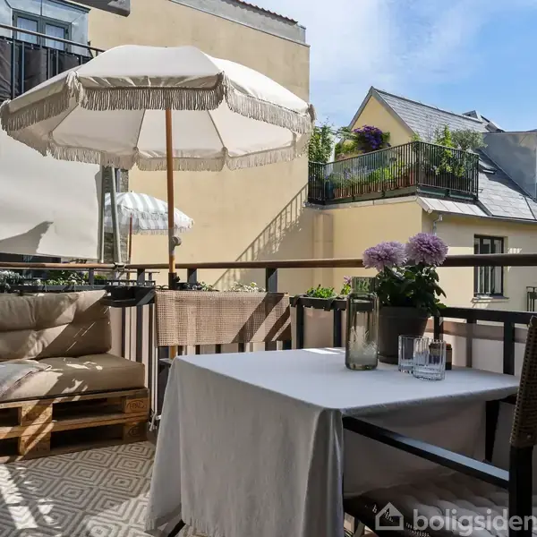 Terrasse med træmøbler; en sofa lavet af paller med hynder står ved siden af et bord med dug. Parasol skygger området omgivet af andre boligbygninger.