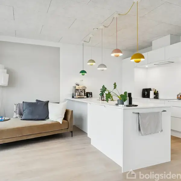 Sofamøbel står i moderne, lyst køkkenmiljø; køkkenø med planter og kaffemaskiner; hængende farverige lamper danner en hyggelig atmosfære. Elegant indretning og store vinduer bringer lys ind i rummet.