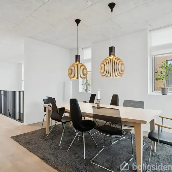 Spisebord med sorte stole står på gråt tæppe i en lys spisestue. To moderne loftslamper hænger over bordet, og planter står i vindueskarmen.