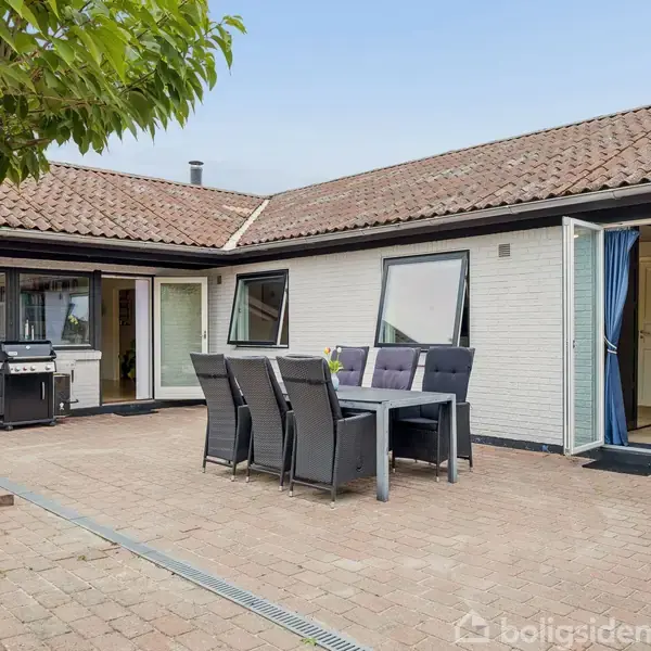 Terrasse med firkantet spisebord og seks stole, grill placeret til venstre ved siden af glasskydedør, omgivet af hvidt murstenshus og have.