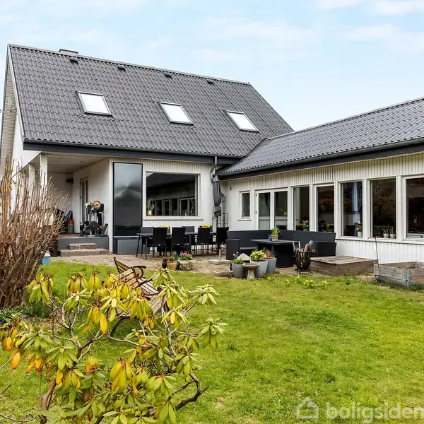 Et moderne, hvidt hus med sort tag har store vinduer og udendørs terrasse. Det står i en grøn have med buske og en græsplæne, omgivet af andre bygninger i baggrunden.
