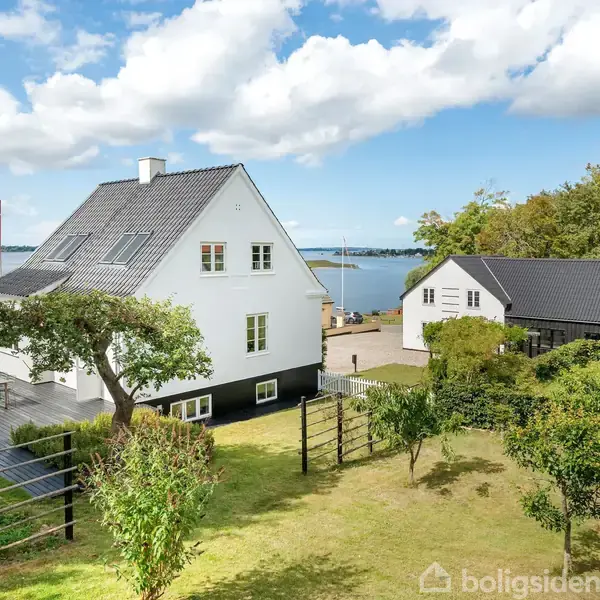 Et hvidt hus står stille i en grøn have med et træ, mens et lyst bådehus ses i baggrunden nær vandet under en blå himmel med få skyer.