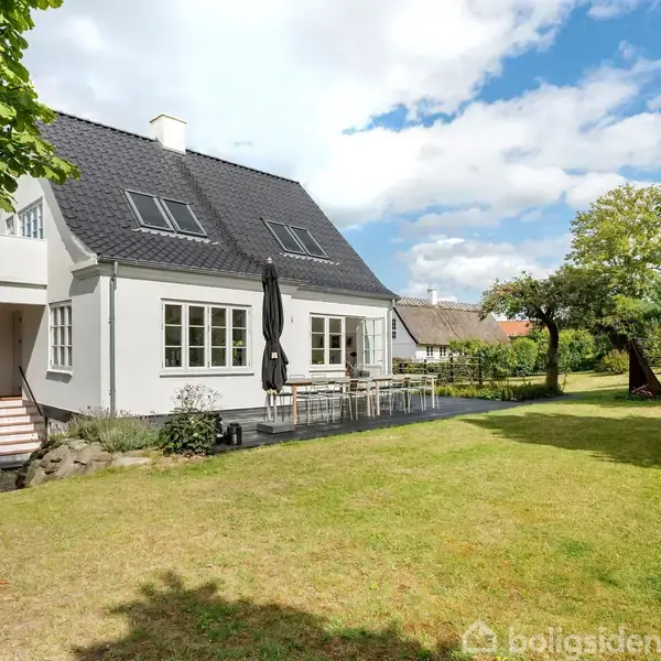 Et lyst rum med loftsbjælker, indeholdende et skrivebord og stol. Store vinduer slipper naturligt lys ind, og der er en sofa samt reoler med bøger.