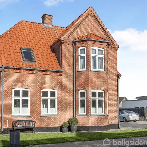 Rødt murstenshus med rødt tegltag står i solbeskinnet gårdhave. Huset har flere hvide vinduer og en bueformet front. Græsplæne og brostensgang omgiver huset.