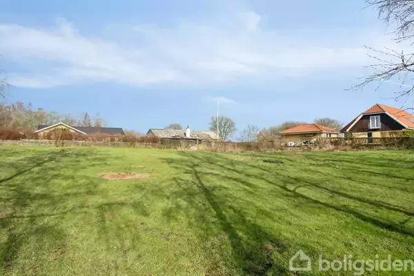 En stor, grøn græsplæne strækker sig mod en række af huse i baggrunden, omgivet af træer og buske under en klar blå himmel.