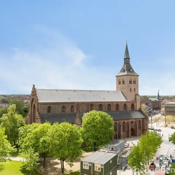 Kirke med et højt spir rejser sig over omkringliggende bygninger og grønne træer. Skyfri himmel i baggrunden og gader med få biler omkring kirken.