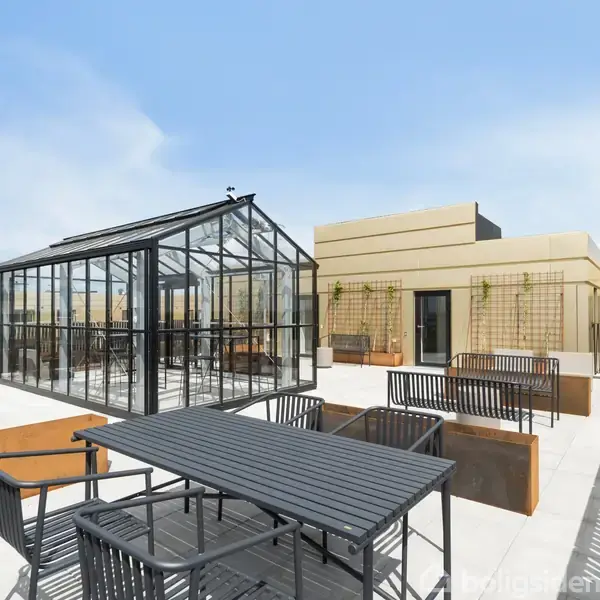 Et sort havebord med stole står på en tagterrasse ved siden af et glashus. Solrigt vejr, moderne bygning i baggrunden. Stabilt rækværk omgiver området. Ingen tekst til stede.